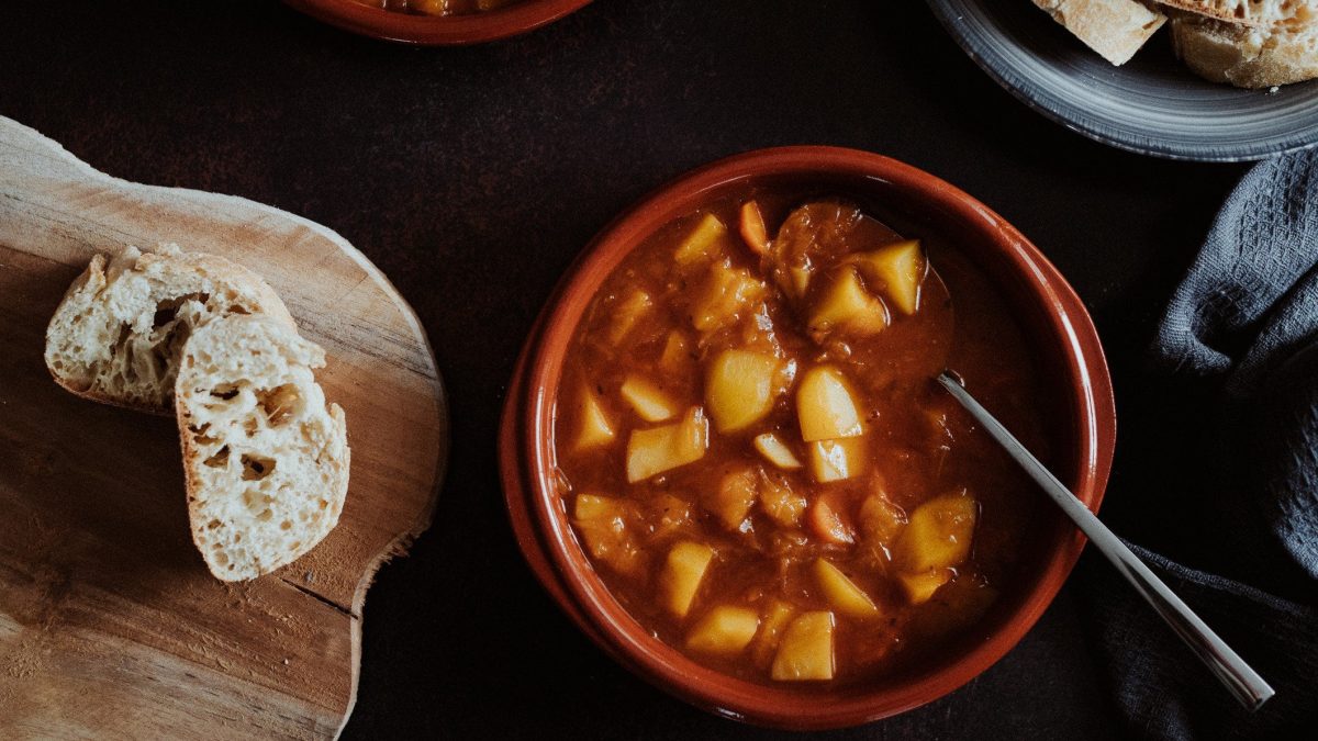 Ein Tontopf mit vegetarischem Kartoffelgulasch in der Draufsicht. Daneben ist ein Brett mit ein paar Scheiben Baguette.