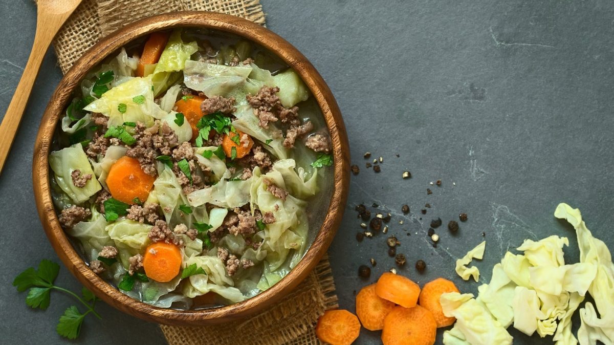 Ein Holzpot mit Weißkohleintopf mit Hackfleisch, Karotten und Kartoffeln, garniert mit Petersilie neben rohen Karotten und Weißkohlstücken, Pfefferkörnern und einem Holzlöffel vor einem grauem Hintergrund, von oben.