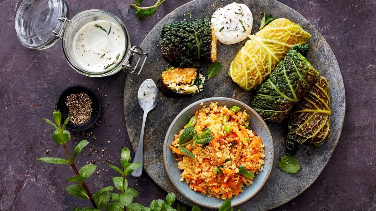Vier Wirsingrouladen mit Couscous gefüllt auf einer runden Steinplatte zusammen mit einem Klacks Joghurt-Soße und einer Schüssel Gmeüse-Couscous serviert, neben einem Glas mit der Soße, einer kleinen Schüssel mit Pfefferkörnern und frischer Minze, vor einem dunklen Hintergrund, von oben.