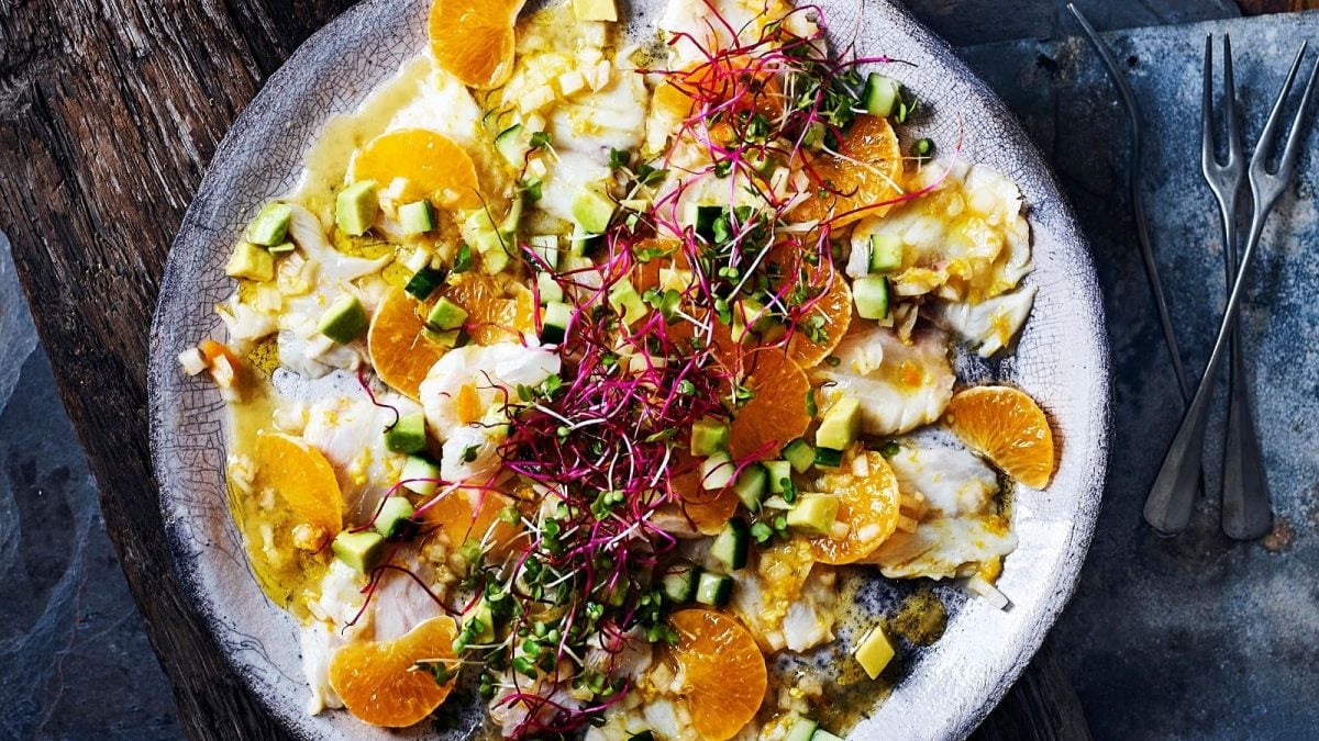 Ein Teller mit Wolfsbarsch-Ceviche mit Mandarinen garniert mit Gurken- und Avocado-Stücken sowie Shisokresse und Radieschensprossen, neben Besteck vor einem Holzhintergrund von oben.