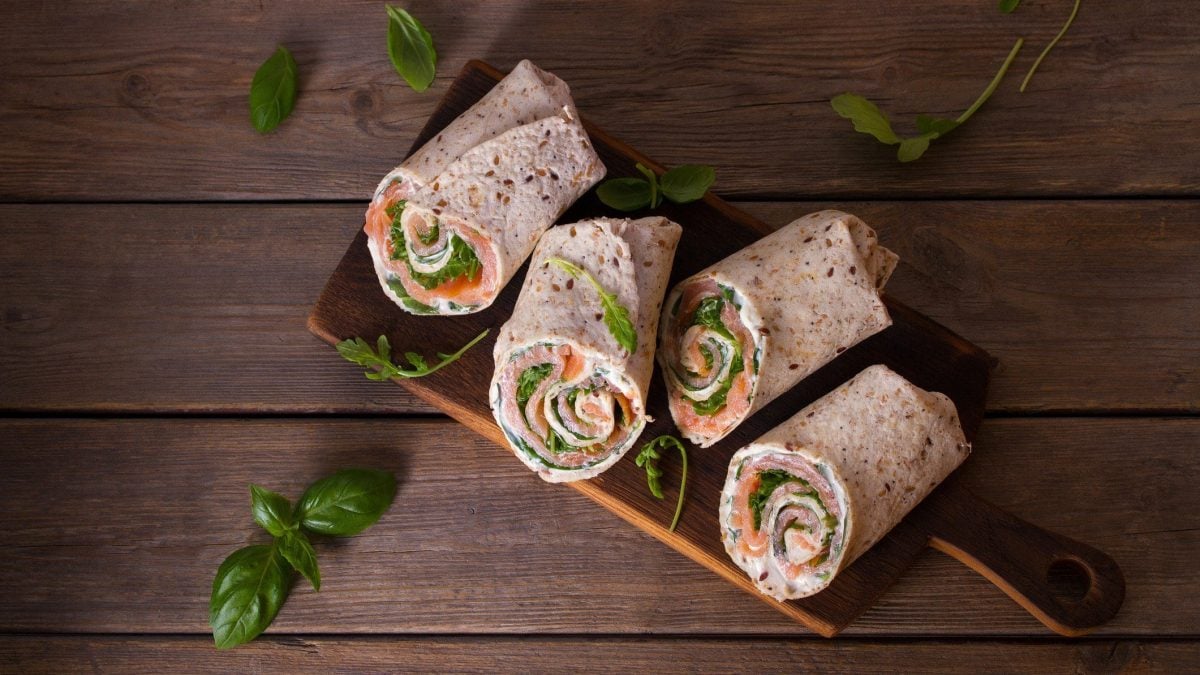 Ein Holzbrett auf einem Holz-Untergrund, auf dem Wraps mit Lachs und Meerrettich liegen. Drumherum ein paar Rucolablätter. Alles in der Draufsicht.