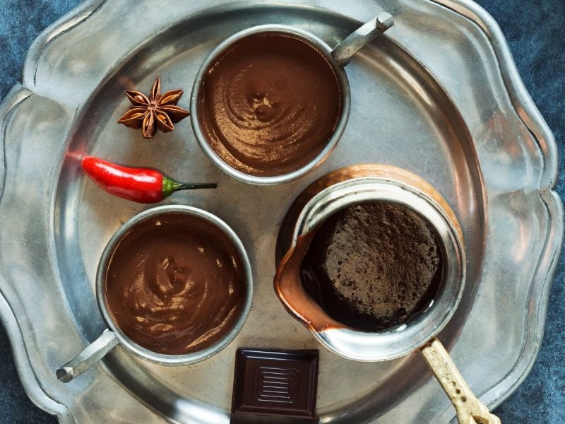 Zwei Tassen gefüllt mit Xocolatl neben einer Kanne, einem Stück zartbitterschokolade, einer Chilischote und einem Stück Sternanis auf einem silbernen Tablett vor einem dunkelblauem Hintergrund, von oben.