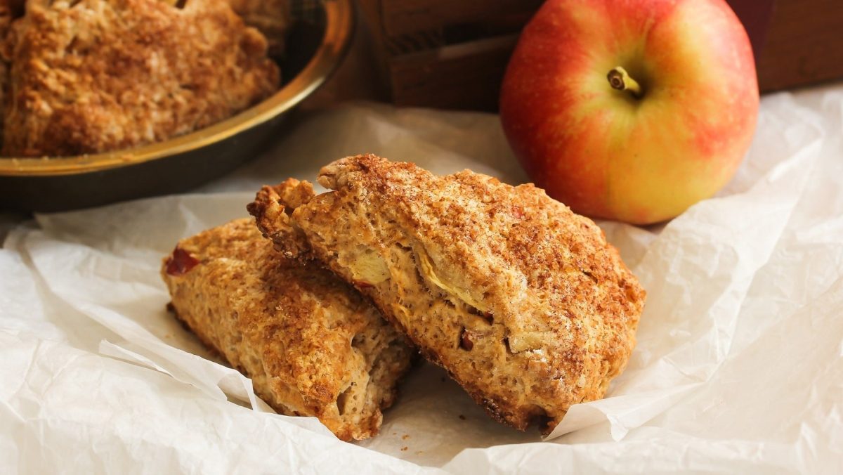 Zwei Apfel-Scones liegen auf gestapelt übereinander auf hellem Backpapier. Im Hintergrund liegt ein Apfel und eine Schüssel mit Scones.