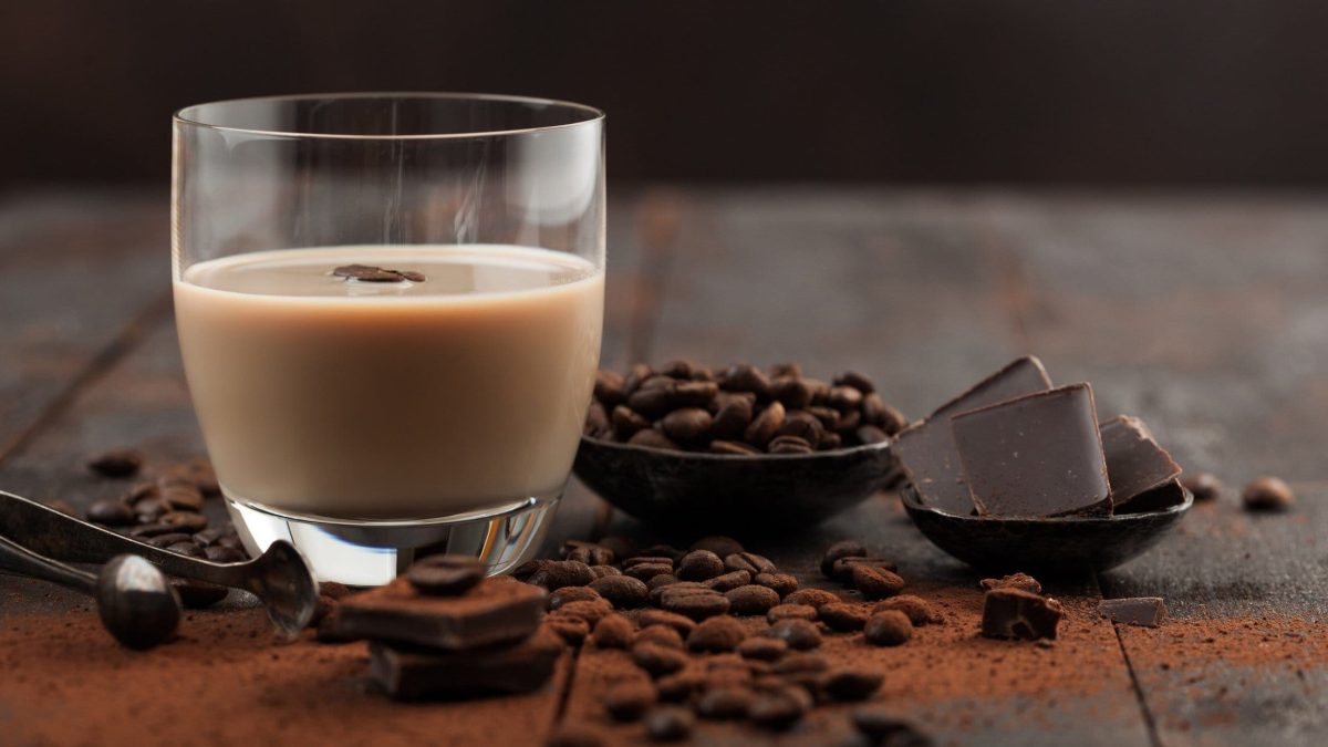 Baileys in einem Glas, umgeben von Schokolade und Kaffeebohnen auf hölzernem Untergrund.
