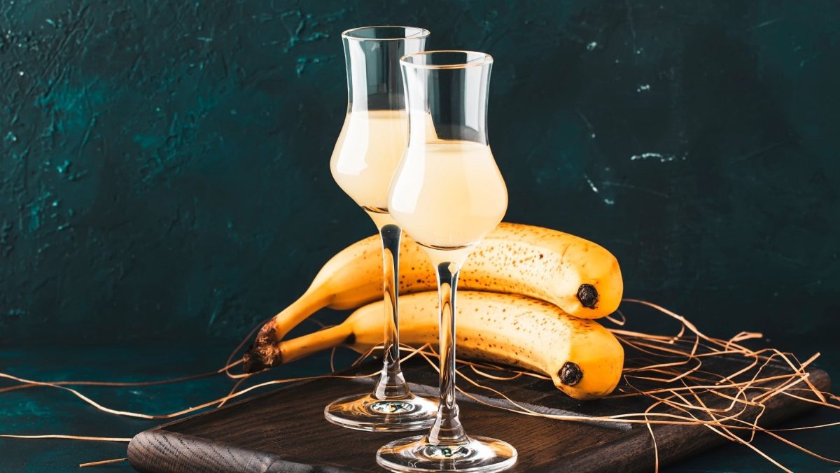 Zwei Gläser mit Bananen-Likör stehen auf einem Holzbrett auf einem dunklen Untergrund. Der Hintergrund ist aus blau-schwarzen Steinen. Hinter den Gläsern liegen zwei Bananen und einige trockene Gräser.