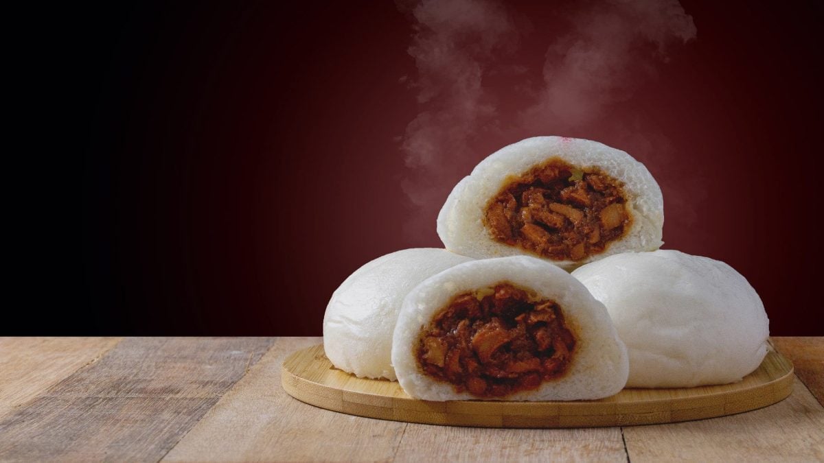 Vier Baozi auf Holzplatte und Holztische. Zwei halbierte lasse die dunkelbraune Char Siu Schweinefleisch-Füllung sichtbar werden. Dampf steigt auf.