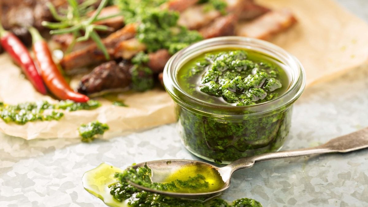 Veganes grünes Pesto in Glas auf Serviette au der Frontalansicht. Davor ein Teelöffel mit Pesto und Olivenöl, das leicht verläuft. Im Hintergrund Kräuter, Chili und Nüsse leicht verschwommen auf Holzbrett angedeutet.