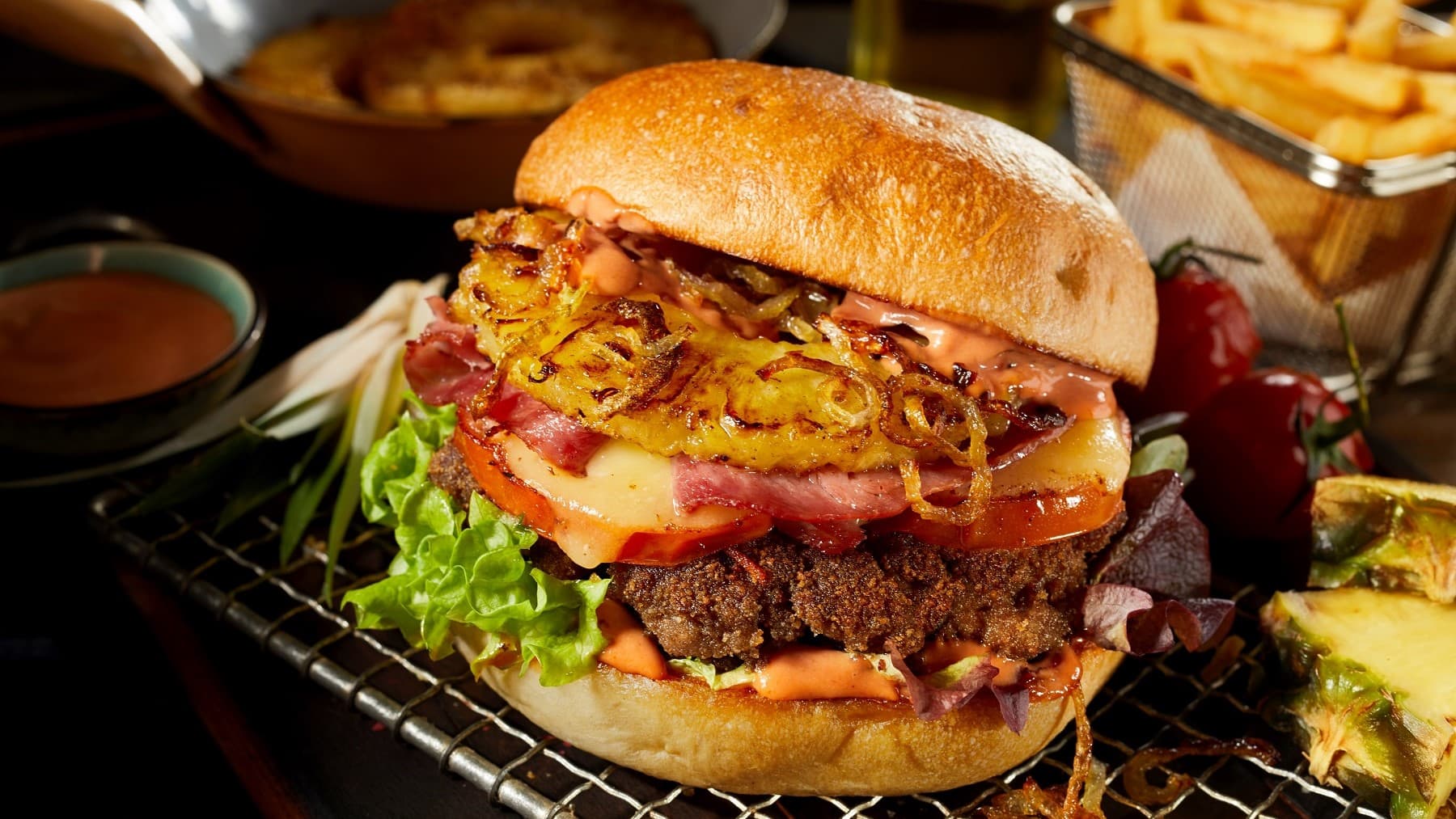 Ein Burger-Hawaii mit Ananas, Speck und gebratenen Zwiebeln liegt auf einem Gitter. Um den Burger herum steht eine Schale mit Coktailsuppe, Ananasstückchen und einige Tomaten. Im Hintergrund liegen weitere Ananasscheiben und eine Portion Pommes.