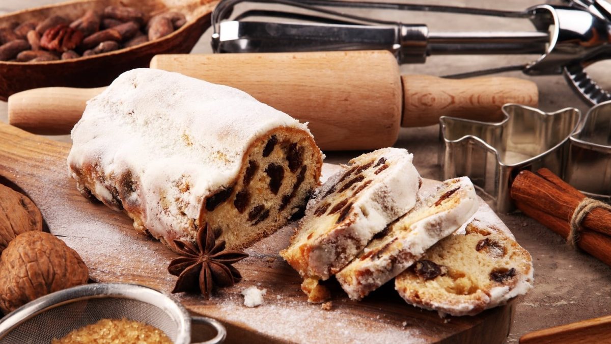 Ein angeschnittener Christstollen steht auf einem Holzbrett umgeben von verschiedenen Backutensilien, Nüssen, Zucker und Zimtstangen. Der Stollen ist verziert mit Puderzucker und angerichtet mit Sternanis.