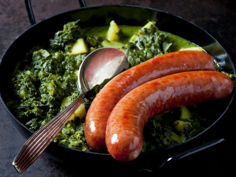 Zwei Knacker auf Grünkohl mit Kartoffelwürfeln und schwarzer Gusspfanne und Esslöffel auf Steinplatte aus der Draufsicht.