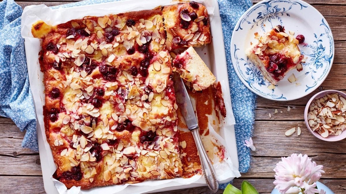 Ein Kiba-Pudding-Butterkuchen in einer Form steht auf einem blau gemusterten Geschirrtuch auf einem braunen Holztisch. Der Kuchen ist bereits angeschnitten und ein Stück steht auf einem blau-weißen Teller mit Blütenmuster. In der Form liegt ein Messer. Rechts neben dem Kuchen steht eine kleine Schüssel mit Mandelblättchen.