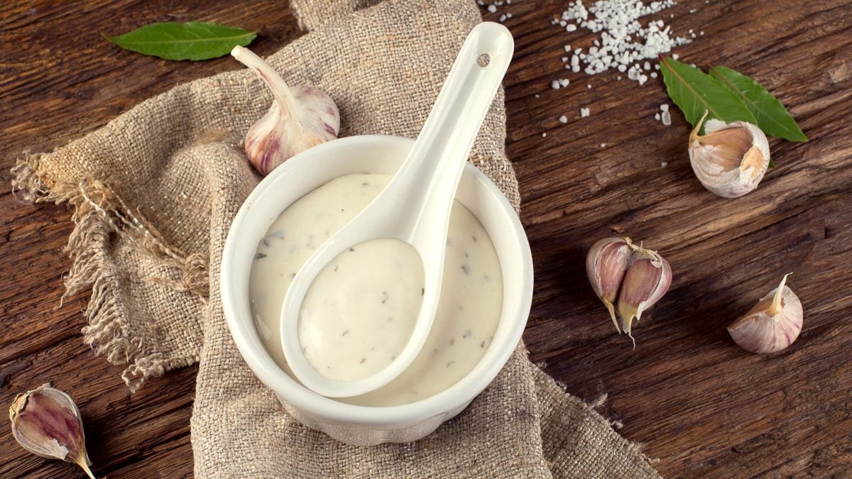 Knoblauchsauce in weißer Schale auf Holzuntergrund. Die Szene ist dekoriert mit einem Leinentuch, Knoblauchzehen und Kräutern.