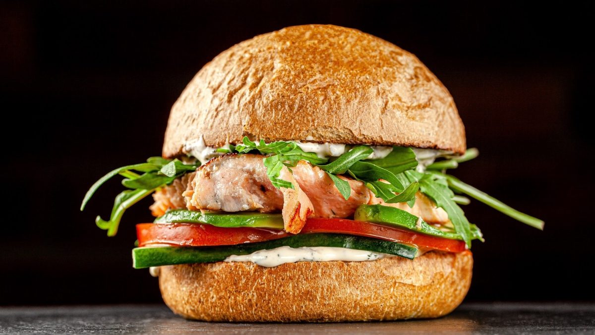 Ein Lachs-Burger mit Avocado, Tomate, Gurke, Remoulade und Rucola steht auf einem glänzenden Untergrund vor einer schwarzen Wand.