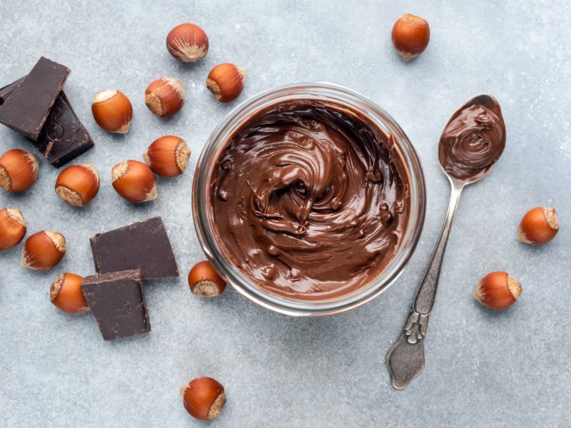 Nuss-Nougat-Creme im Glas umgeben von den zutaten auf hellblauem Untergrund