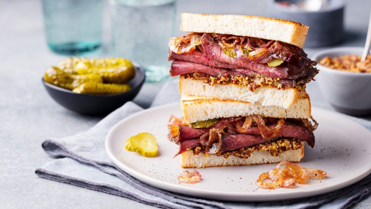 Pastrami-Sandwich mit Senf, karamellisierten Zwiebeln und Gurken auf einem weißen Teller, umgeben von Zutaten.