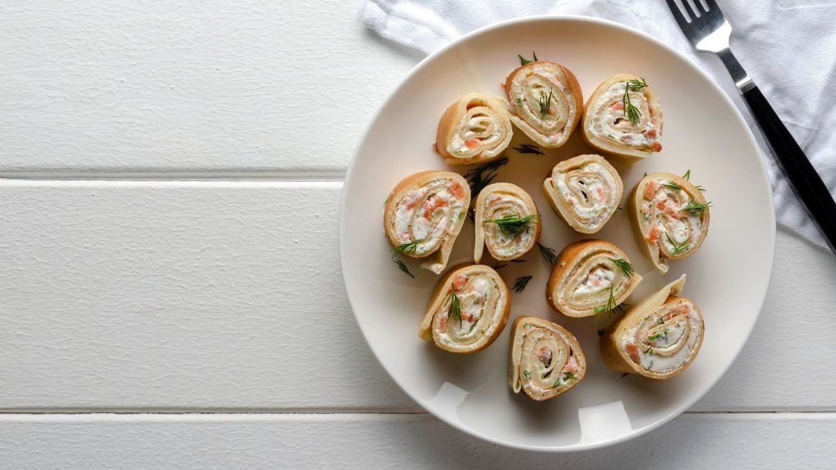 Pfankuchenröllchen mit Lachs auf weißem Teller drapiert auf weißém Holz-Untergrund.