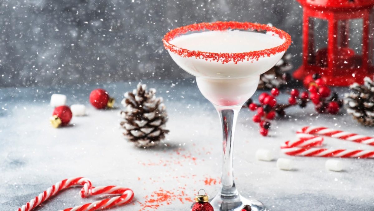 Ein Schneeflocken-Daiquiri in einem mit Zuckerperlen verzierten Glas auf einem Untergrund aus Stein. Um das Glas herum liegen Zuckerstangen, Kienäpfel und Christbaumkugeln. Künstlicher Schnee rieselt im Hintergrund.