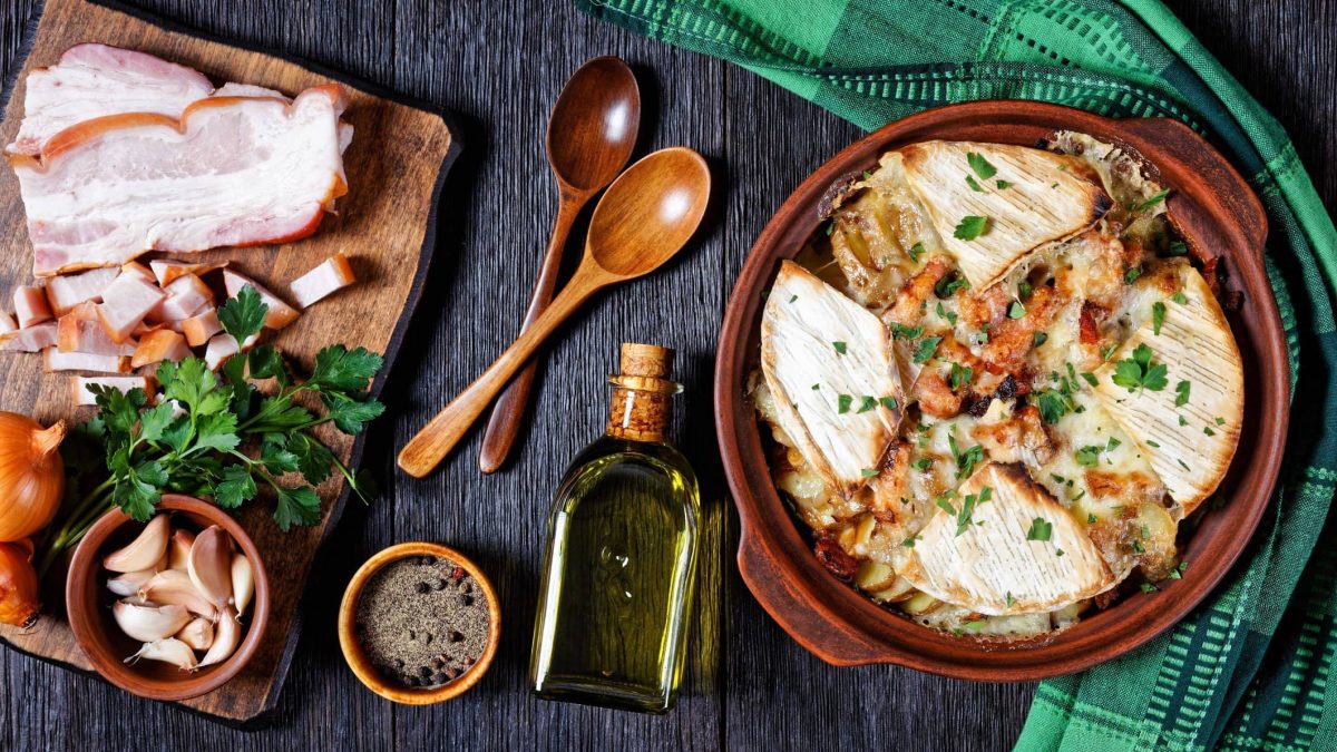 Tartiflette mit Reblochon und Speck in Auflaufform auf dunklem Holz, umgeben von verschiedenen Zutaten.
