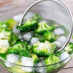In einer Schüssel aus Glas sind viele Eiswürfel und Brokkoli-Röschen. In der Schüssel ist auch ein Küchensieb aus Metall.