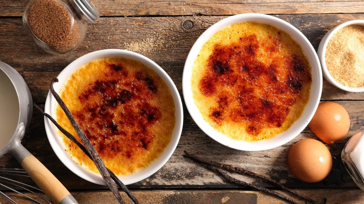 Zwei weiße Förmchen mit Amelies Crème Brûlée mit extra krosser Karamellschicht gefüllt, neben Vanilleschoten, Eiern, Zucker und Milch, vor einem Holzhintergrund, von oben.