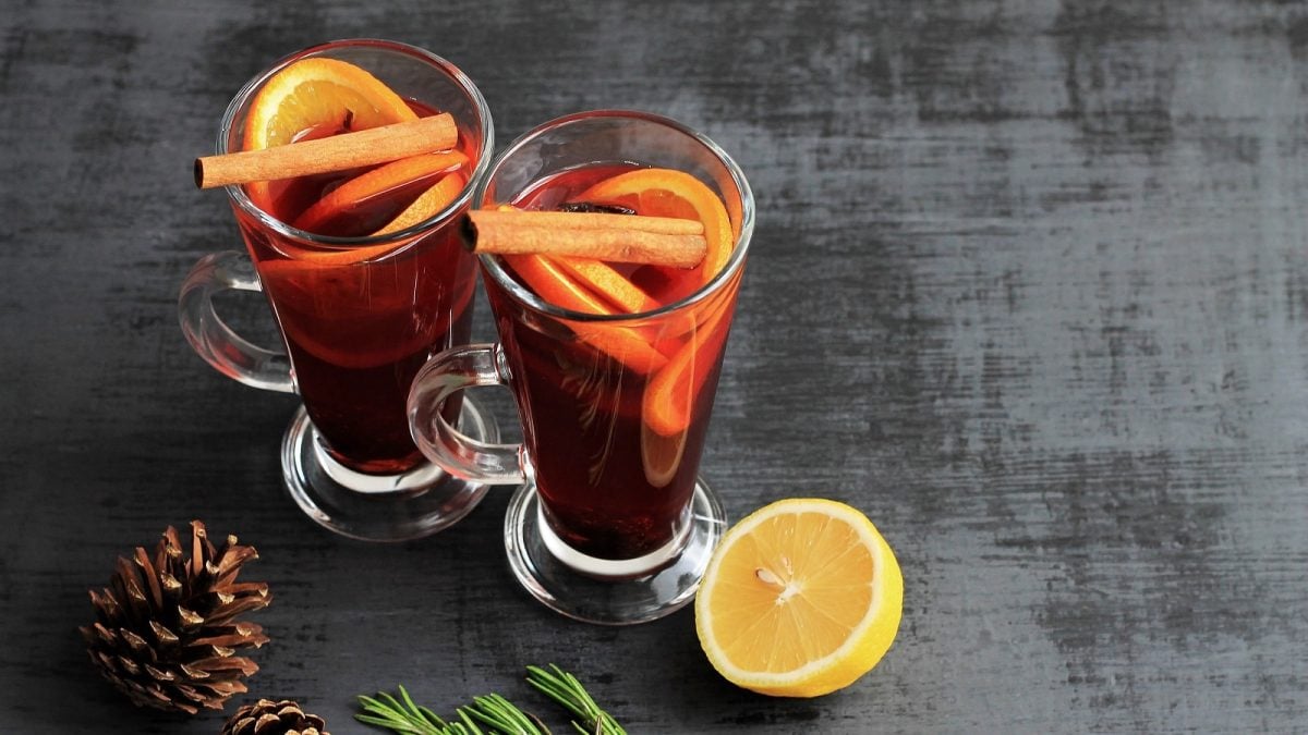 Zwei hohe Gläser gefüllt mit dem Apfel-Holunder-Orangen Punsch mit viel Gewürzen, garniert mit Orangenscheiben und Zimtstangen, neben einer halben Zitrone, einem Tannenzapfen und einem Tannenzweig.