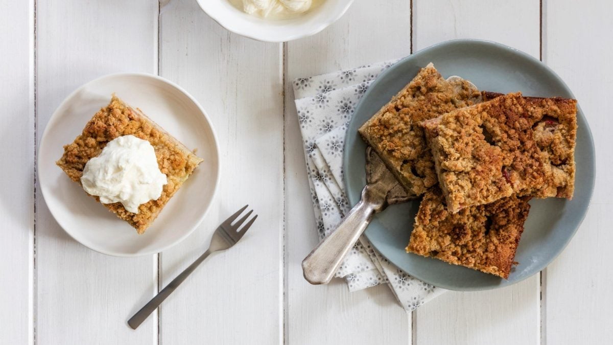 In der Draufsicht sieht man zwei Teller mit einigen Stücken Apfel-Schmand-Kuchen auf weißem Holz. Außerdem Servietten und eine Kuchengabel.