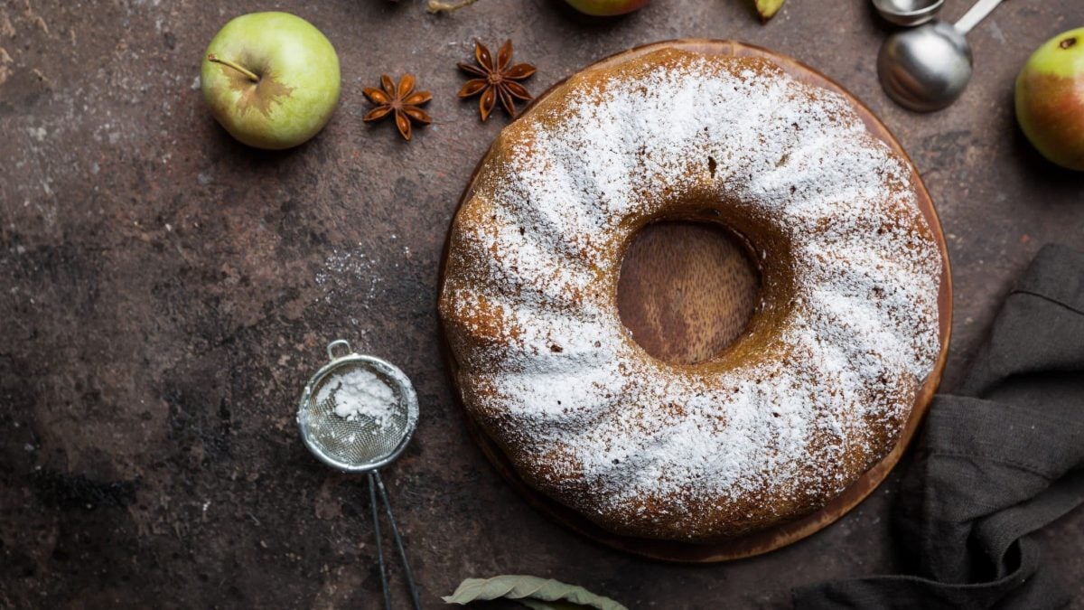 Ein Apfelkuchen mit Kardamom, ein Topfkuchen, in der Draufsicht. Daneben liegen Äpfel, ein Sieb mit Puderzucker und Sternanis.