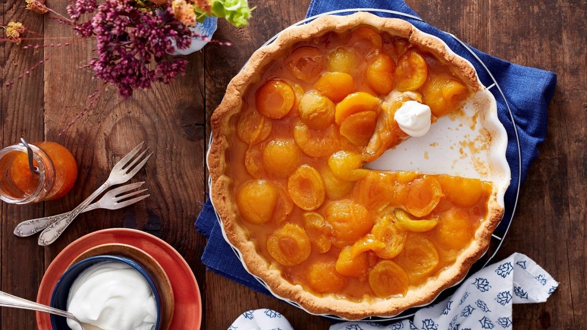 Eine Tarte-Form gefüllt mit der Aprikosen-Tarte mit Espresso-Creme bei der ein Stück fehlt und die mit etwas Schlagsahne garniert ist, neben einer Schüssel Schlagsahne, einem Glas Konfitüre, Gabeln und Blumen vor einem Holzhintergrund, von oben.
