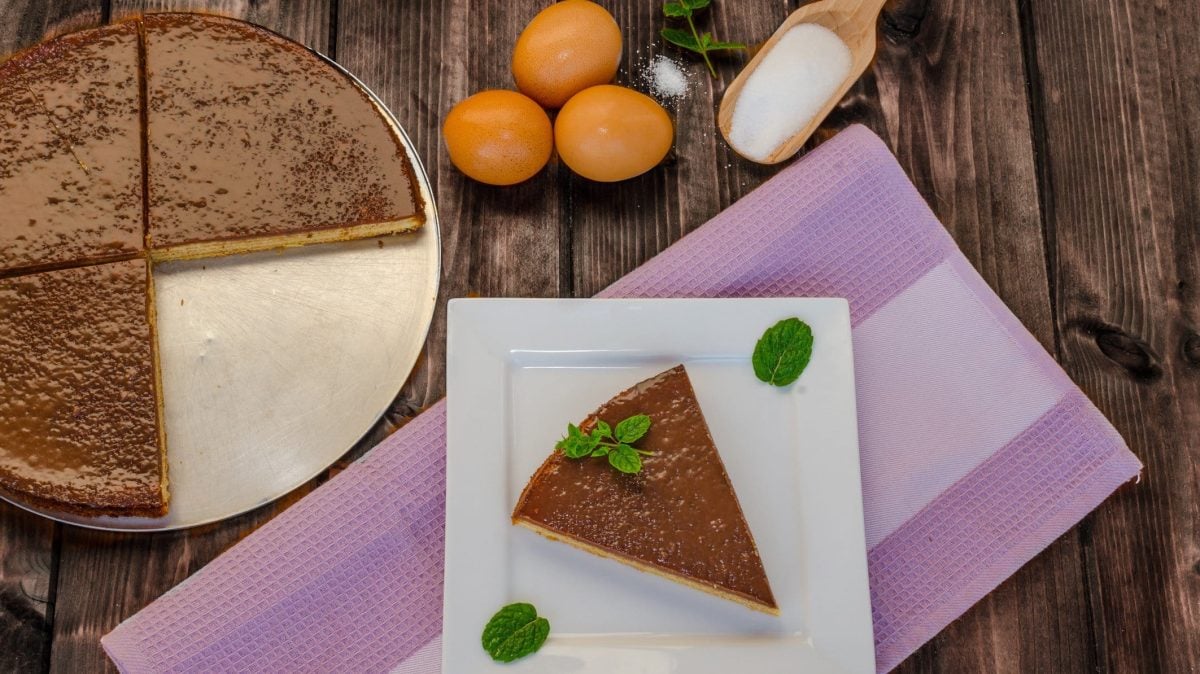 Ein angeschnittener Baumkuchen auf rustikalem Holzuntergrund in der Draufsicht. Außerdem Eier und ein Holzlöffel Zucker.