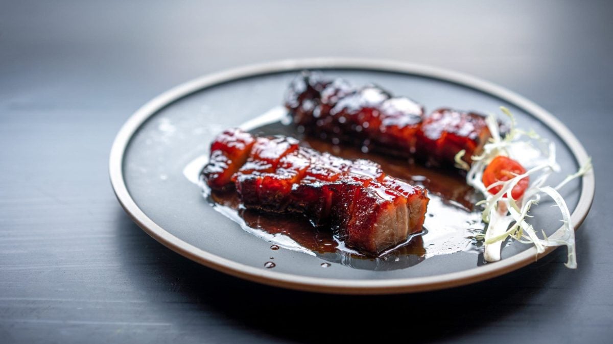 Aufgeschnittenes Schweinefleisch Char Siu aus der Frontalsicht. Rotbräunliche Färbung durch Röstung auf grauem Teller und grauem Untergrund. Rechts aufgeschnittener weißer Rettich..