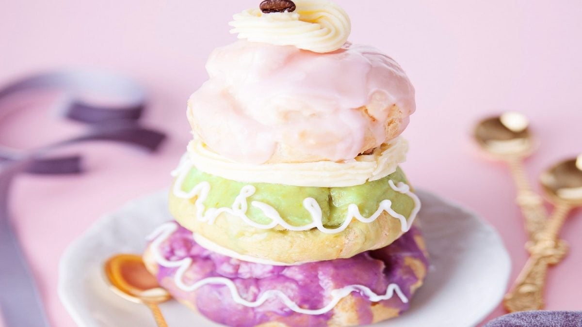 Eine Courtesan au Chocolat wie im Grand Budapest Hotel: Drei aufeinandergestapelte, mit Schokoladen-Creme gefüllte und mit farbigem Zuckergüss und Frosting verzierte Brandteig-Bäööe auf einem weißen Teller neben goldenen Löffeln vor einem rosa Hintergrund.