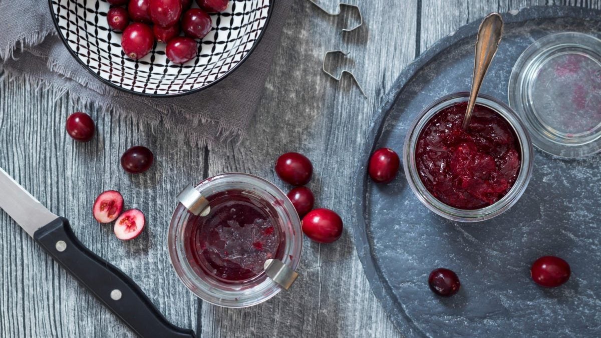 In der Draufsicht sieht man Weckgläser mit Cranberry-Soße, außerdem frische Cranberries und ein Messer auf Holzplanken.