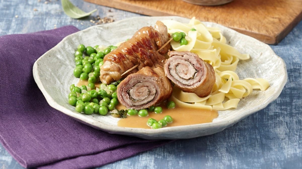 Auf einem Teller liegt eine Roulade. Sie ist aus zartem Kalbsfleisch und wird mit Salbei gewürzt. Dazu gibt es Erbsen und Nudeln
