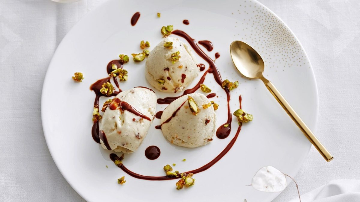 Drei Stück Stollen-Mousse mit Mokkasoße und Pistazienkrokant garniert neben einem goldenen Löffel auf einem weißen Teller, vor einem hellen Hintergrund.