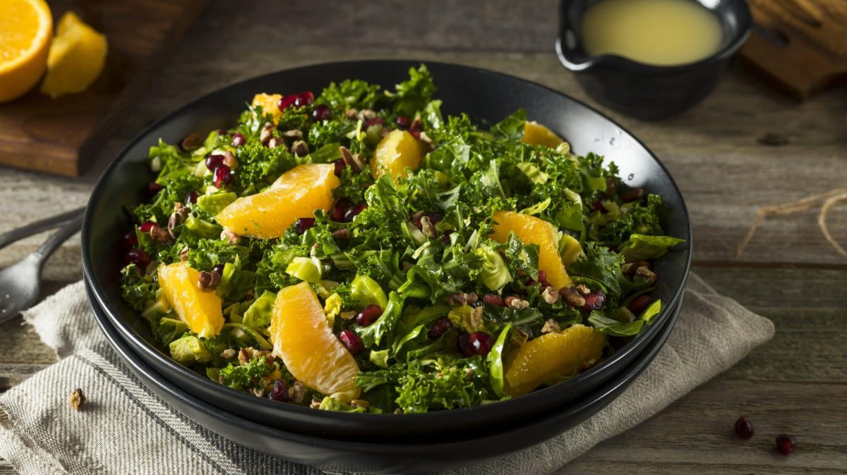 In einer runden Schale ist Grünkohlsalat mit Orange und Walnüssen Auf dem Tisch sind zwei Orangenhälften zu sehen.
