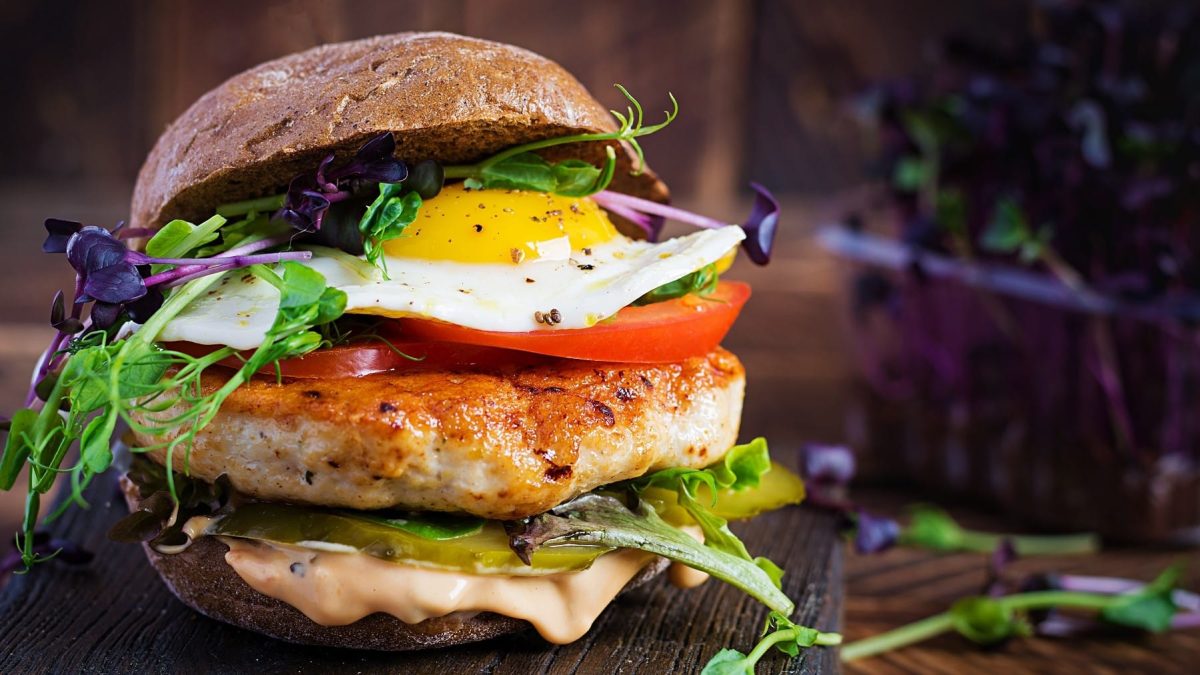 Ein Hähnchen-Burger mit Spiegelei steht auf einem dunklen Holzbrett auf einem dunklen Untergrund. Im Hintergrund steht eine Schale mit Salat.