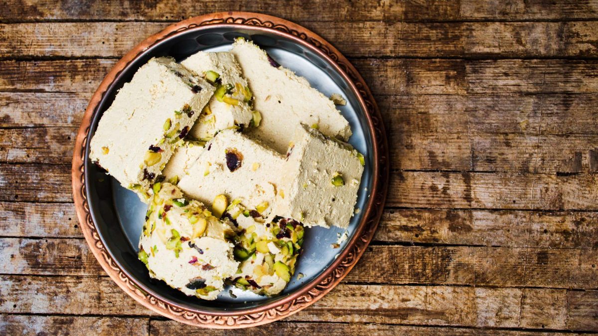 Auf einem Holzuntergrund sieht man in der Draufsicht einen runden Teller mit Halva mit Pistazien.