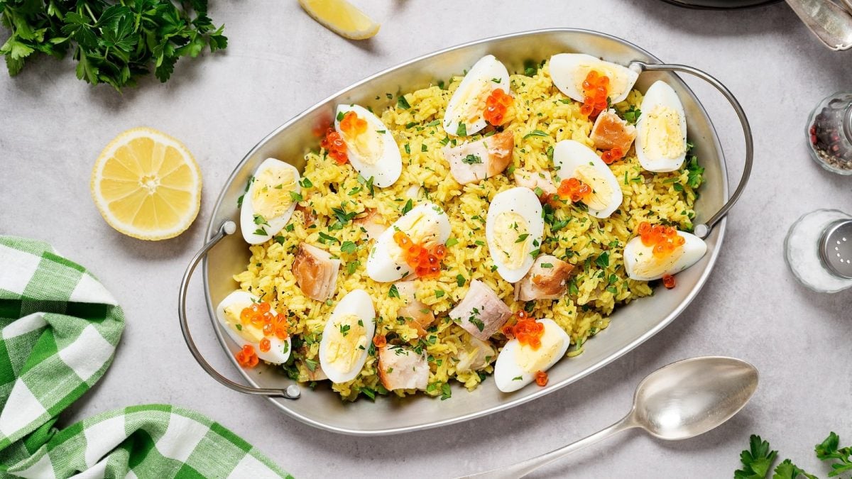 Eine Servier-Schüssel angerichtet mit Kedgeree, einem indischen Gericht mit britischer Adaption, garniert mit rotem Kaviar und Petersilie, neben einem Löffel, einer halben Zitrone, Salz, Pfeffer, Petersilie und einem Tuch, vor grauem Hintergrund, von oben.