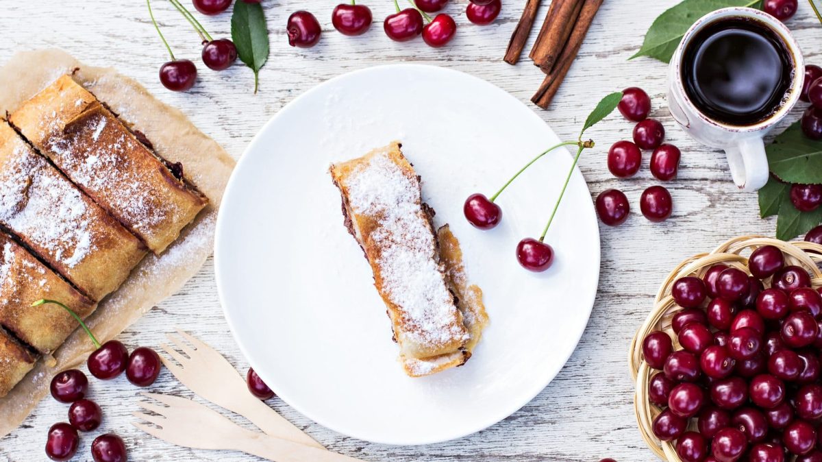Draufsicht von oben: Auf einem weißen Tisch liegen Kirschen. Auf einem weißen Teller liegen mehrere Stücke Kirschstrudel.