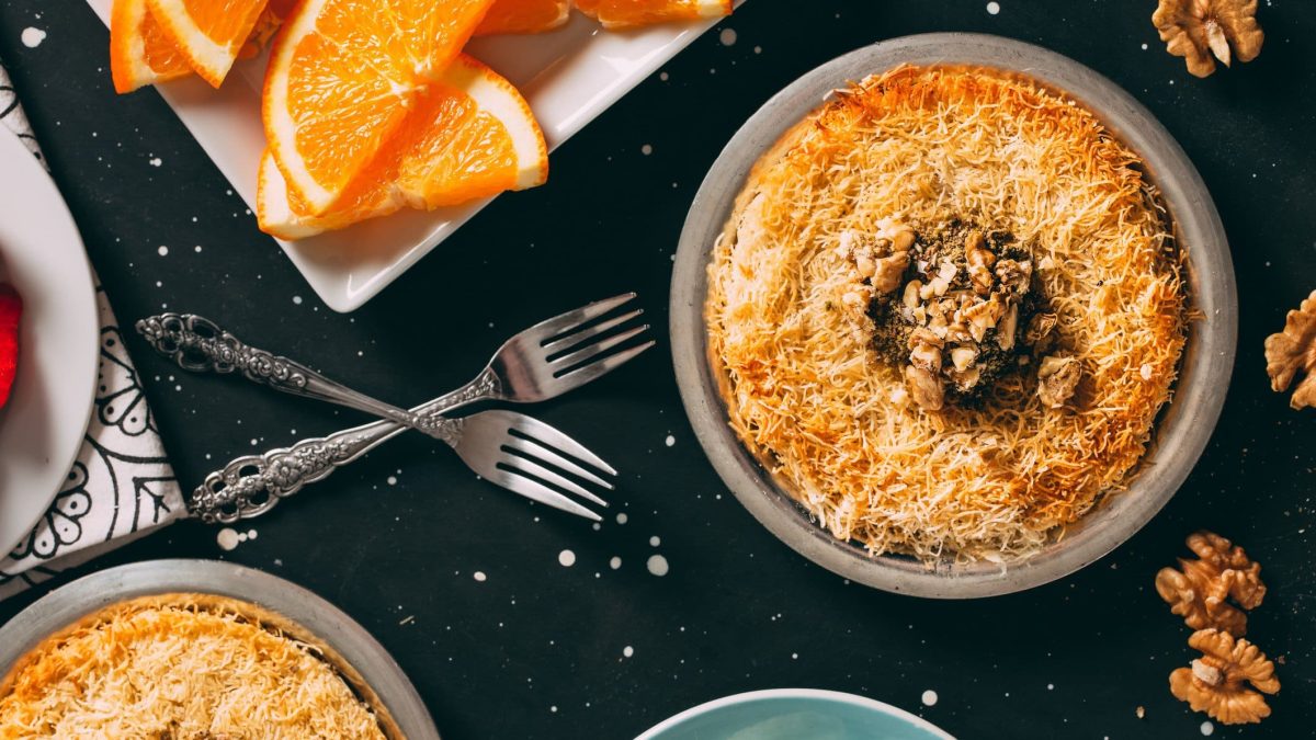 Eine Schale mit Knafeh auf dunklem Untergrund in der Draufsicht. Drumherum liegen Kuchengabeln, Orangenscheiben und Walnüsse.