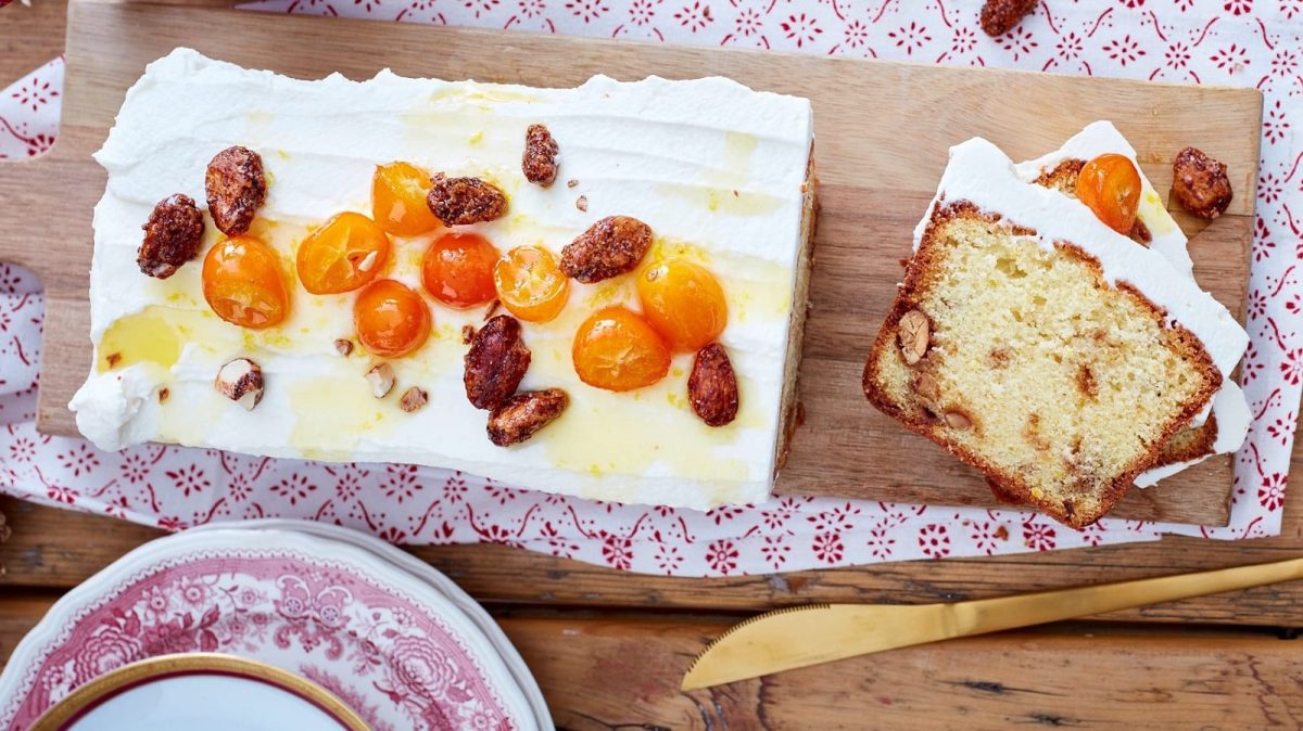 Ein Holzbrett auf dem der Kuchen mit gebrannten mandeln und Kumquats liegt, angeschnitten mit zwei Stücken daneben