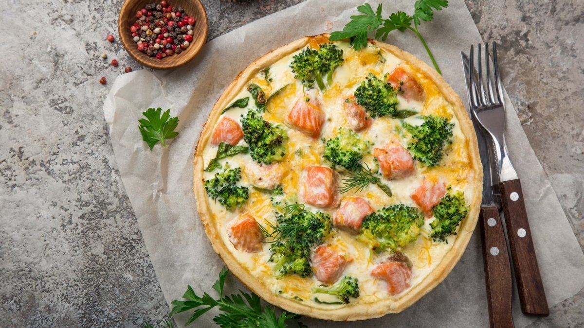 In der Draufsicht: Eine Brokkoli-Lachs Quiche ist in einer runden Form. Daneben liegen Messer und Gabel. Auch eine Schale mit Pfefferkörnern ist zu sehen.