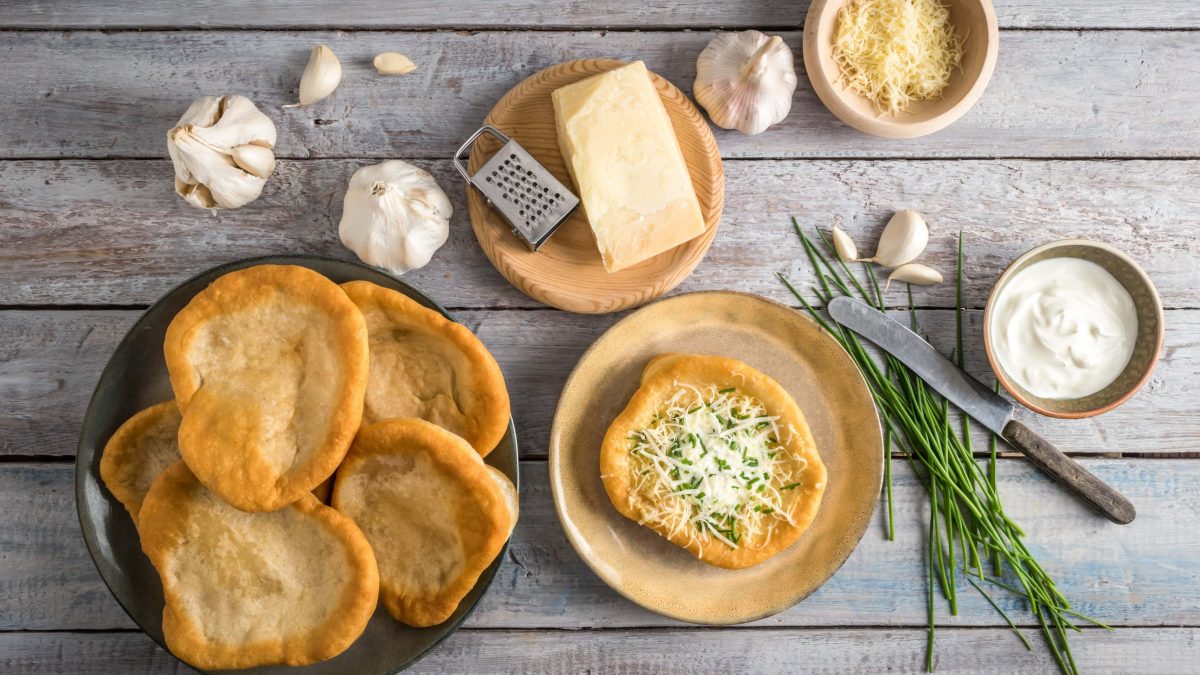 In der Draufsicht sieht man einen Teller mit Langos, daneben ein weiterer Teller mit einem belegten Langos mit Käse. Drumherum sind Zutaten wie Knoblauch und Schmand zu sehen.