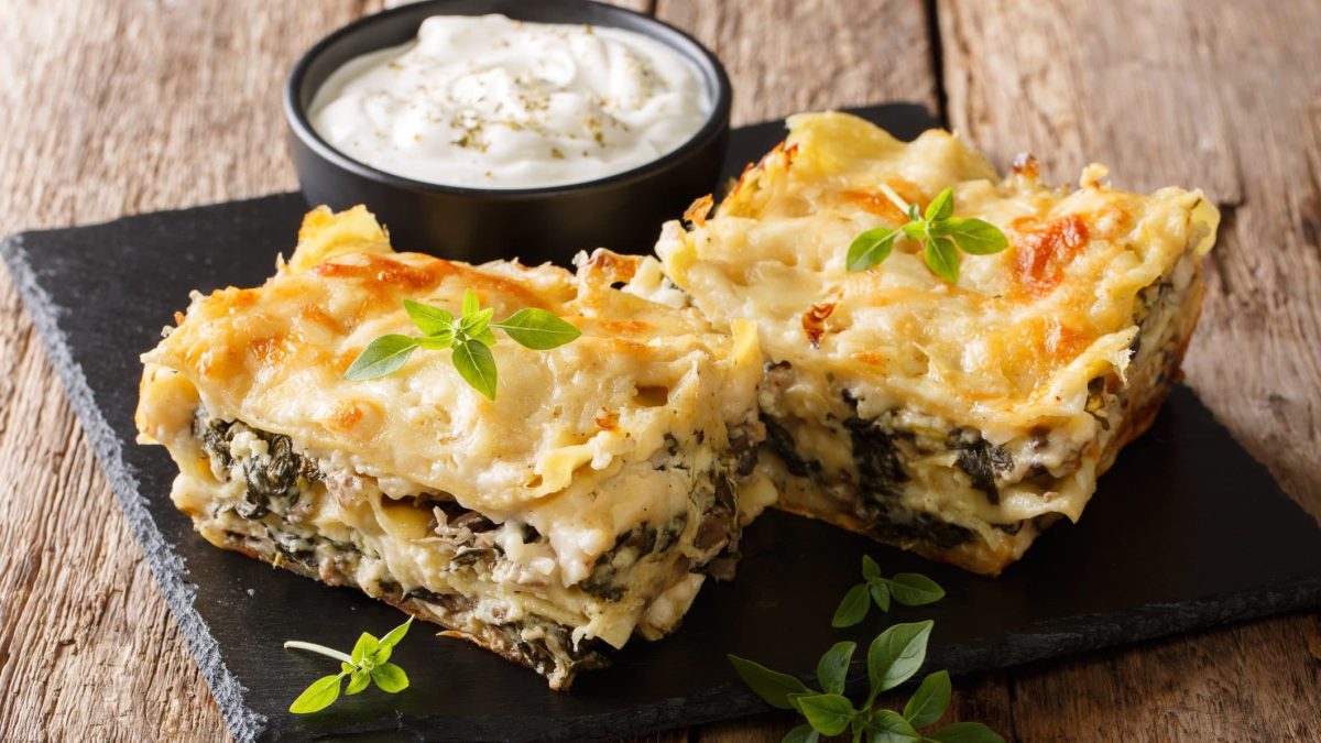 Auf einer schwarzen Schieferplatte liegen zwei Stücke Lasagne mit Spinat und Pilzen, außerdem steht darauf eine Schale mit einem weißen Dip.