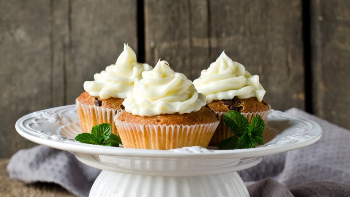 Auf einem Kuchenständer sehen drei Macadamia-Cupcakes mit Frosting vor einer Holzwand.