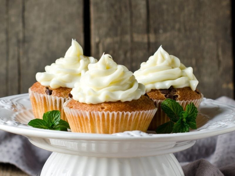Auf einem Kuchenständer sehen drei Macadamia-Cupcakes mit Frosting vor einer Holzwand.