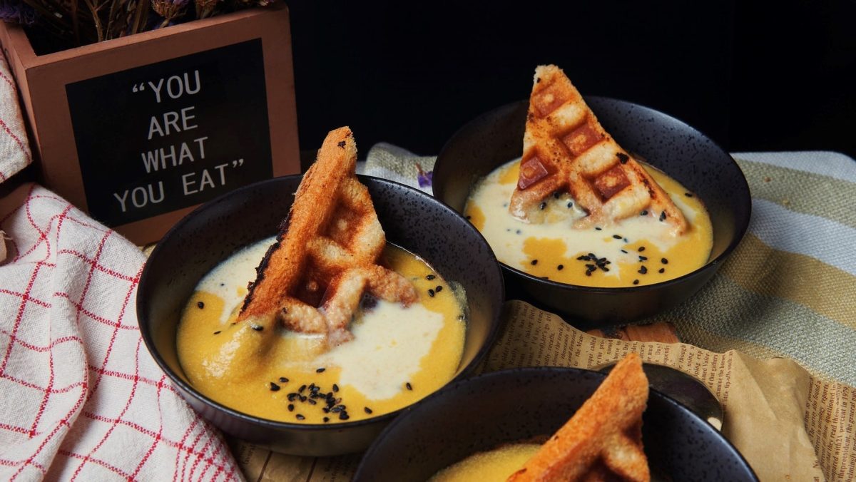 Maissuppe mit Waffeln in zwei schwarzen Schüsseln. Die Suppen sind angerichtet mit Crème fraîche und Sesamsamen. Due Schüsseln stehen auf verschiedenen Geschirrhandtüchern. In der linken oberen Ecke steht ein Schild auf dem steht "You are what you eat"