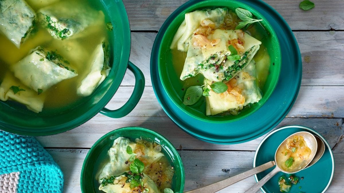 Ein grüner Topf und zwei kleinere Schüsseln mit Portionen der Maultaschen mit Tofu und Grünkohl neben einem kleinen Teller mit Löffeln und einem blauen gehäckelten Untersetzter, vor einem Holzhintergrund, von oben.