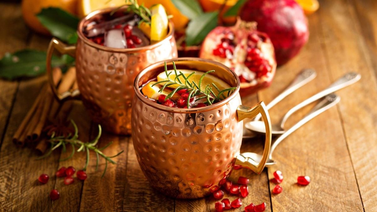 Auf einem Tisch stehen zwei Messingbesser in denen ein Getränk ist. Es ist mit Zitronenscheiben und ROsmarin dekoriert. Auf dem Tisch liegen Granatapfelkerne, sowie ein halber Granatapfel.