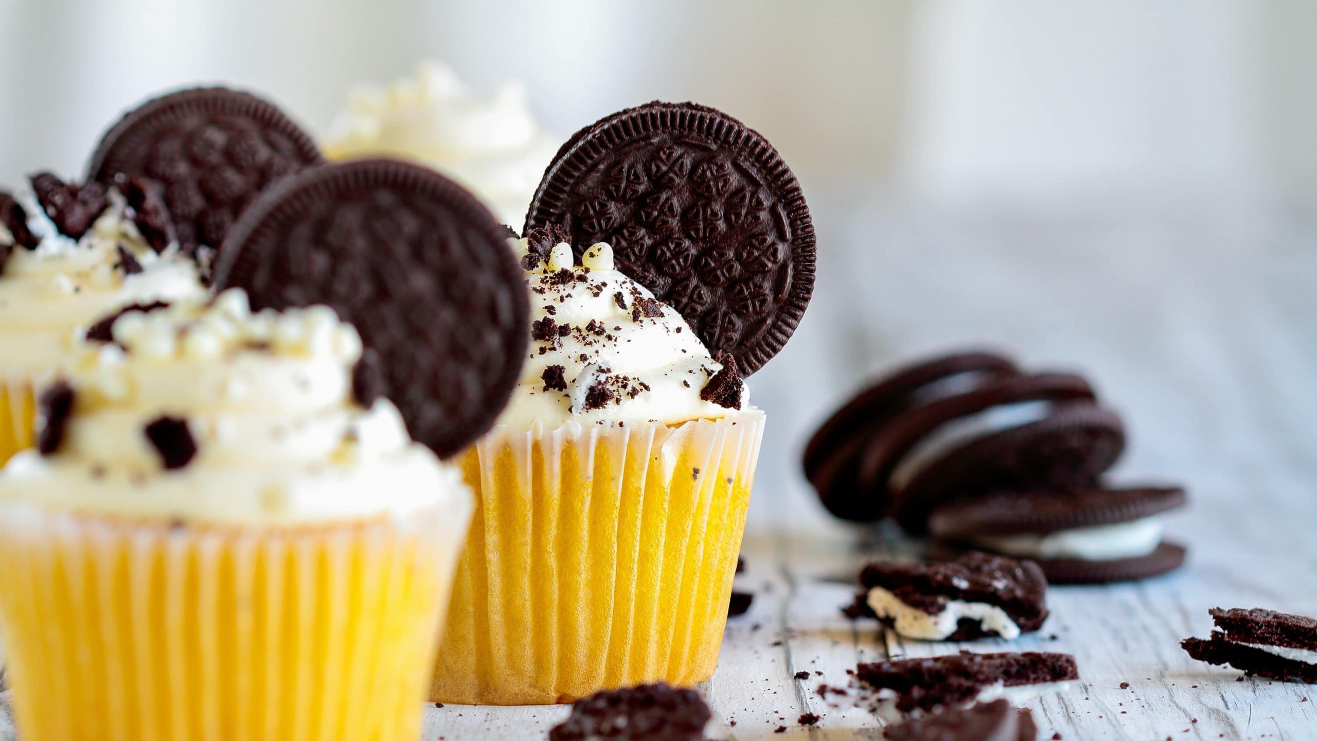 Drei Oreo-Cupcakes in gelben Muffinförmchen auf hellem Holz, daneben liegen ein paar zerbröselte Kekse.